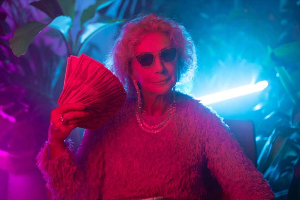 Older lady sunglasses fanning herself with cash and showered in neon light.