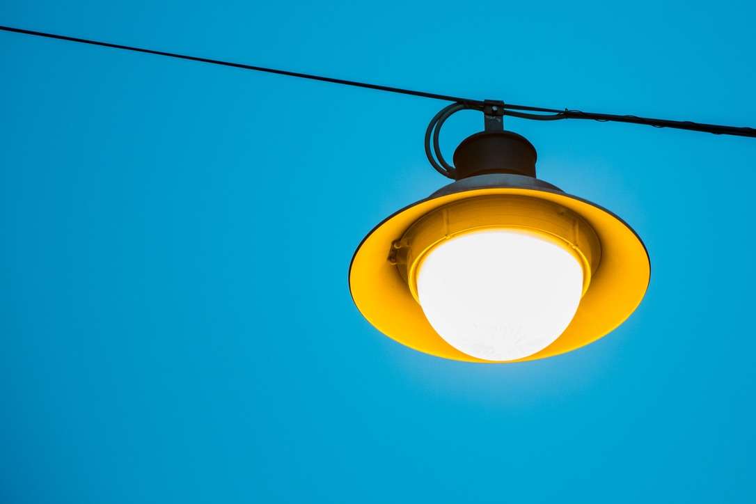 Lit yellow street light on wire in front of a blue sky.