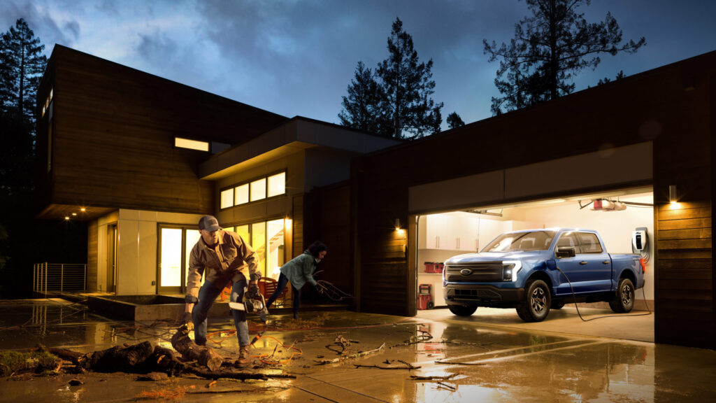 2022 Ford F-150 Lightning Pro electrifying a home with its battery during a grid power outage. The homeowners are cleaning up debris the driveway. Pre-production model with available features shown.