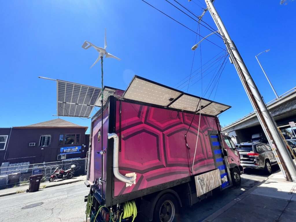 Purple painted moving truck parked on a city street with solar panels covering the roof and hanging over the edges plus a wind turbine attached to the back.