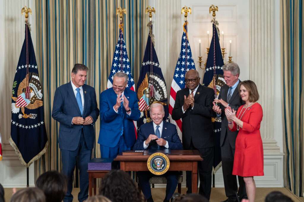 Joe Biden signing the Inflation Reduction Act in the White House, seated in front of 5 clapping US Senators and Representives.