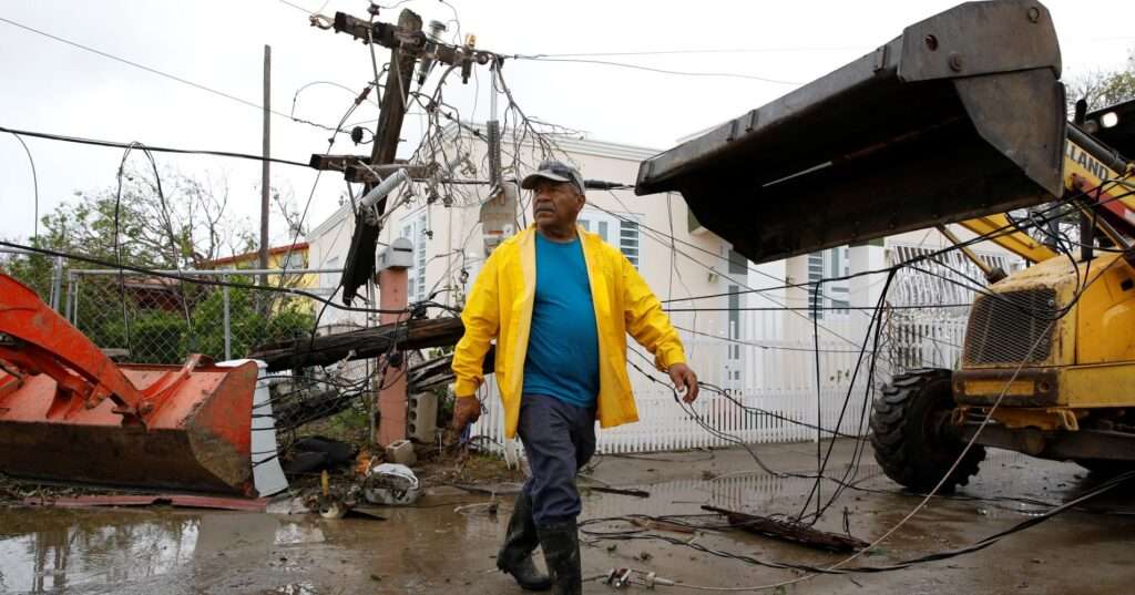 Puerto Rican man walking through a street in front of 2 bulldozers, a broken chainlink fence and many powerlines.