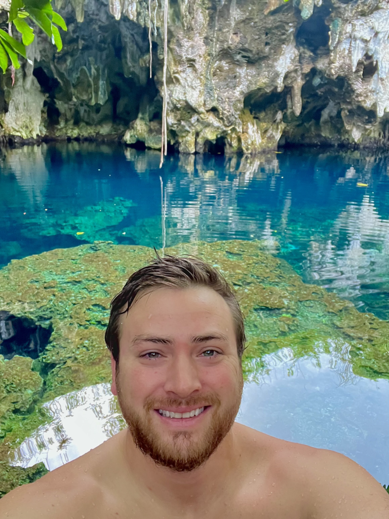 Selfie of Alexander Boom from the shoulders up in front of emerald blue swimming hole on the island of Zanzibar.