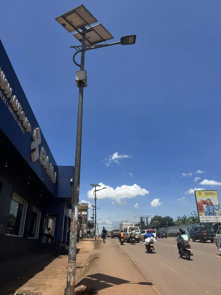 Street lamps with 2 large solar panels attached to their tops line a busy road in Kampala, while the midday sun shines down on them.