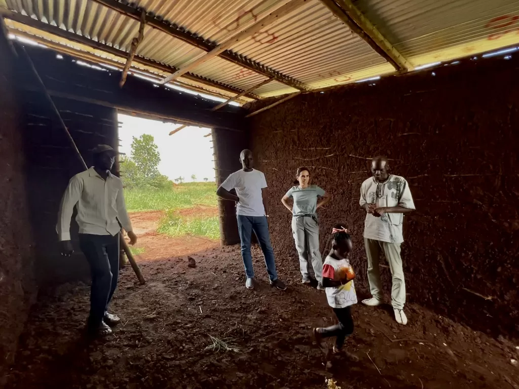 5 people standing in a mud-walled dirt-floored, and an aluminum-roofed room that's under construction. Ben Male is in the room watching his 6-year-old daughter walk in front.