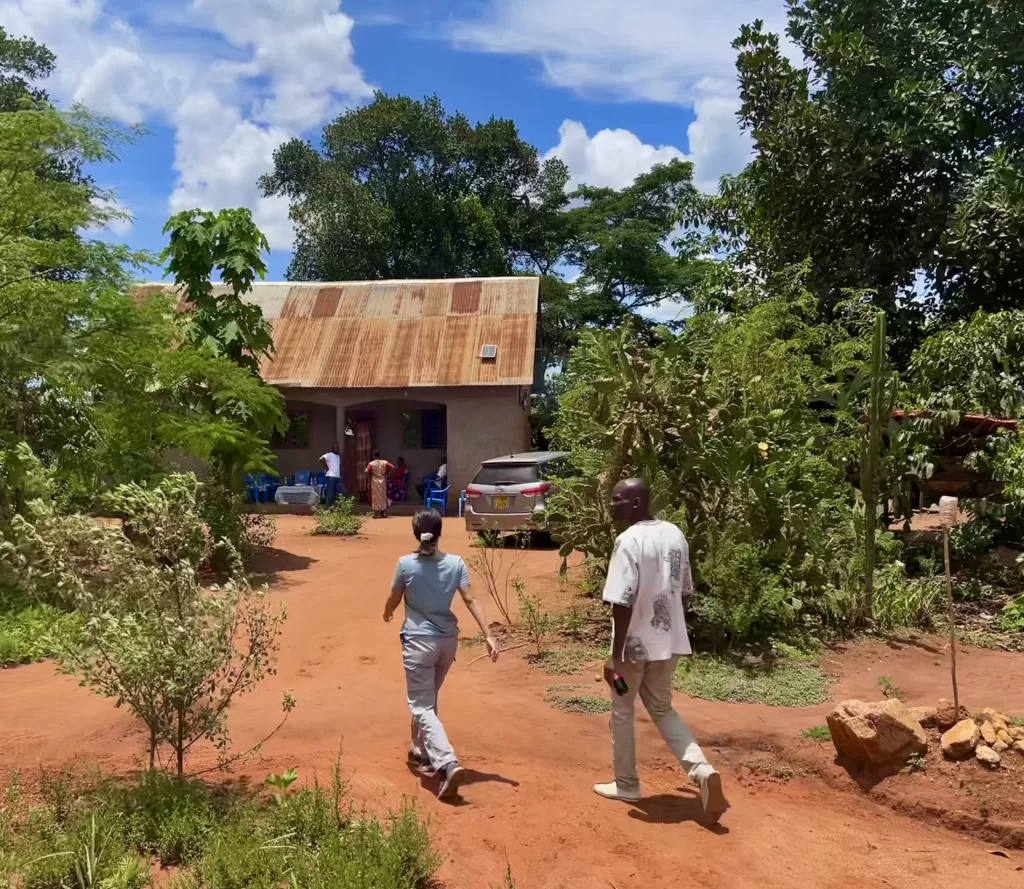 A man and a woman walk up a dirt path to a home surrounded by plants with a group of people talking on the front porch.