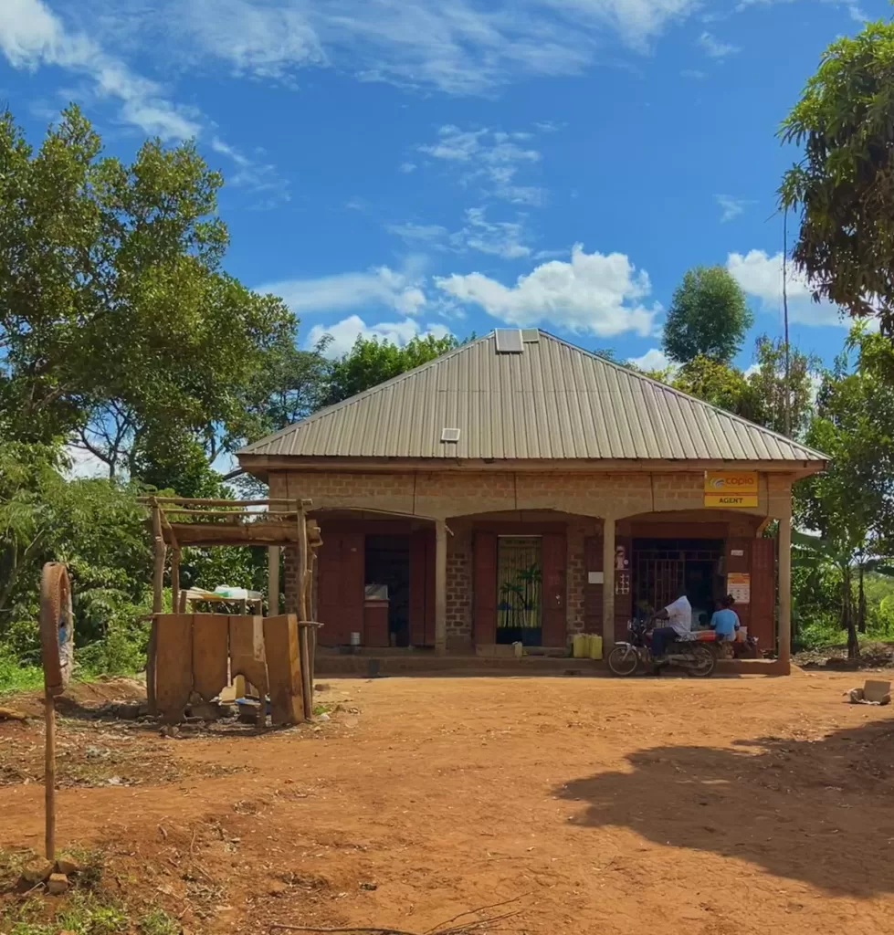 Metal-roofed brick store with a solar panel on top in the Ugandan countryside under a blue sky. A man with a motorcycle pulled up front while a girl sits in the store's front porch.