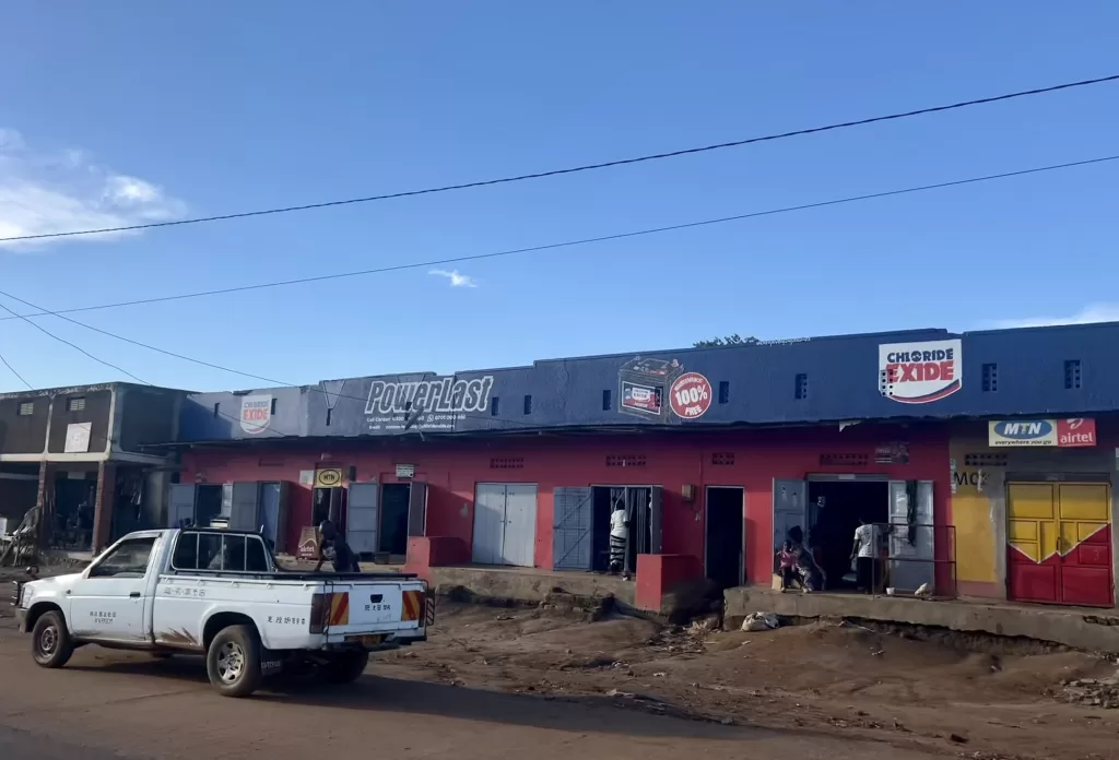 Roadside storefront of a Ugandan energy and battery provider Chloride Exide. The shop is on a dirt bank with a pickup truck out front and few people going into the shop.