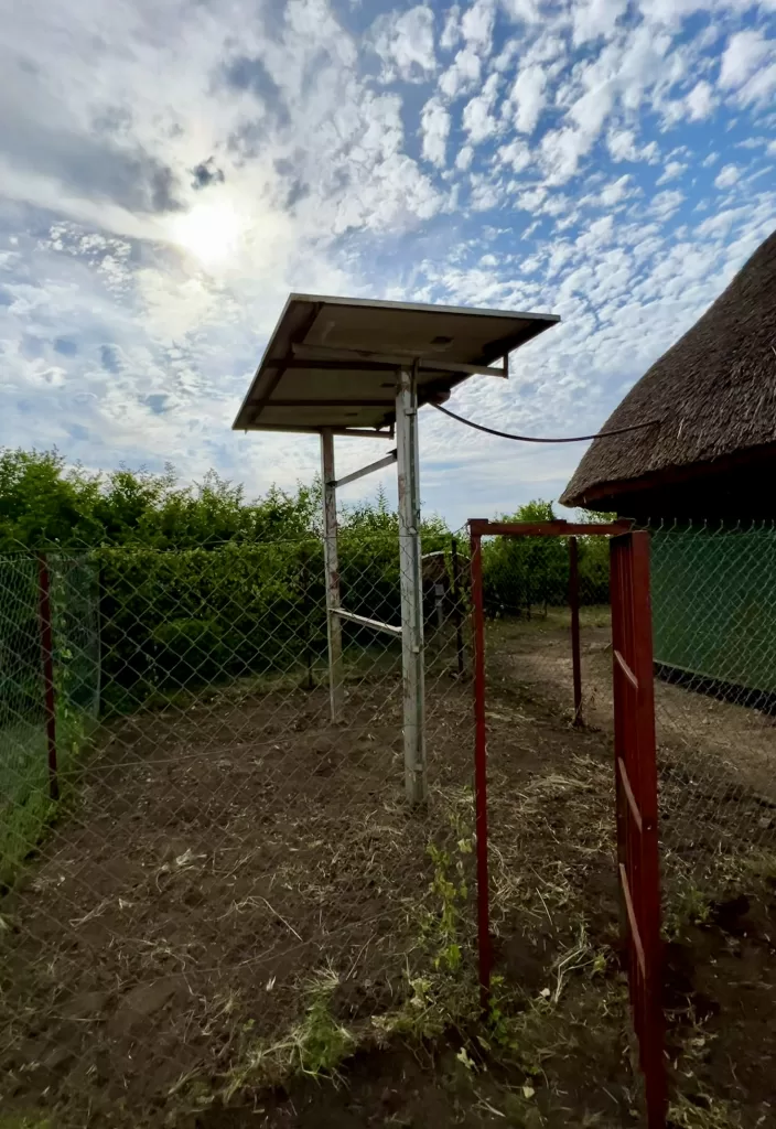 2 large solar panels attached to a platform and connected to a thatched roof building. Solar panels are surrounded by a fence.