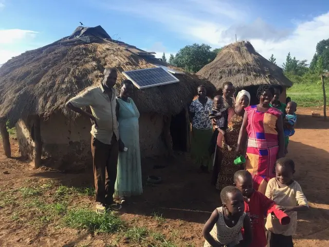 Two thatched roof homes in Kidepo Uganda with their residents (6 women, 5 children, 1 man) standing out front and smiling for the camera. One of the huts is powered by solar energy from the panels on its roof.