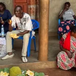 7 adults and 2 young girls eating fruit and drinking soda on the front porch of a farmhouse in Uganda on a Sunny afternoon.