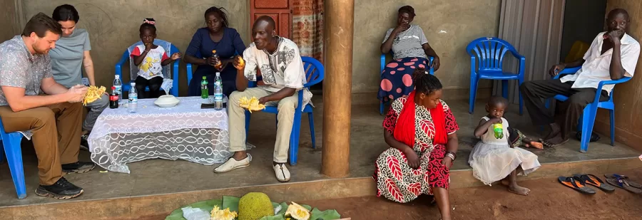 7 adults and 2 young girls eating fruit and drinking soda on the front porch of a farmhouse in Uganda on a Sunny afternoon.