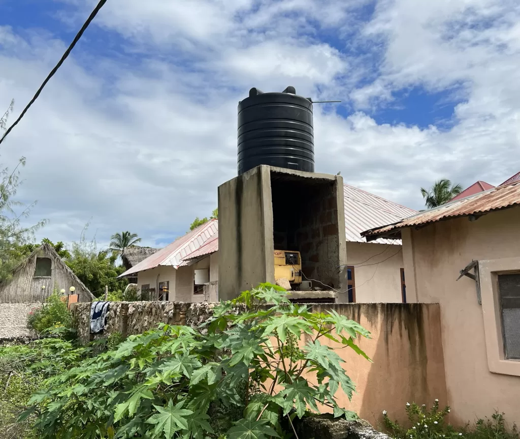 Yellow gas generator on a platform belwo an elevated water tank and connected with wires to a house in Paje, Zanzibar, Tanzania on a sunny afternoon.