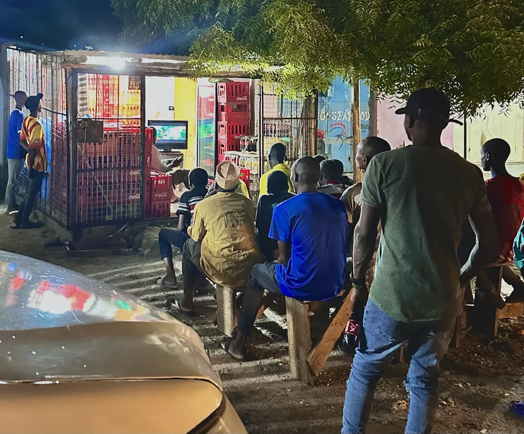 A group of men and boys sitting on wooden benches watching a TV in a store at night in the town of Paje, Zanzibar, Tanzania.