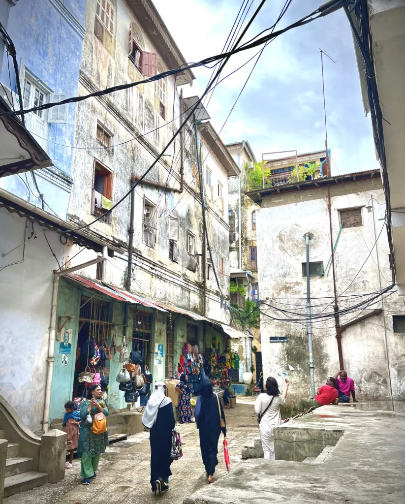 Women in headscarves walking through a narrow powerline-strewn street in Stone Town, Zanzibar, Tanzania as 2 men lounge on a nearby stoop.