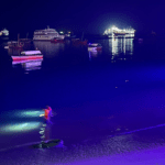 Ocean meets that sand at night in Stone Town Zanzibar. Two people with flash lights walk in the shallow water as people sit at a dockside restaurant and boats float offshore.