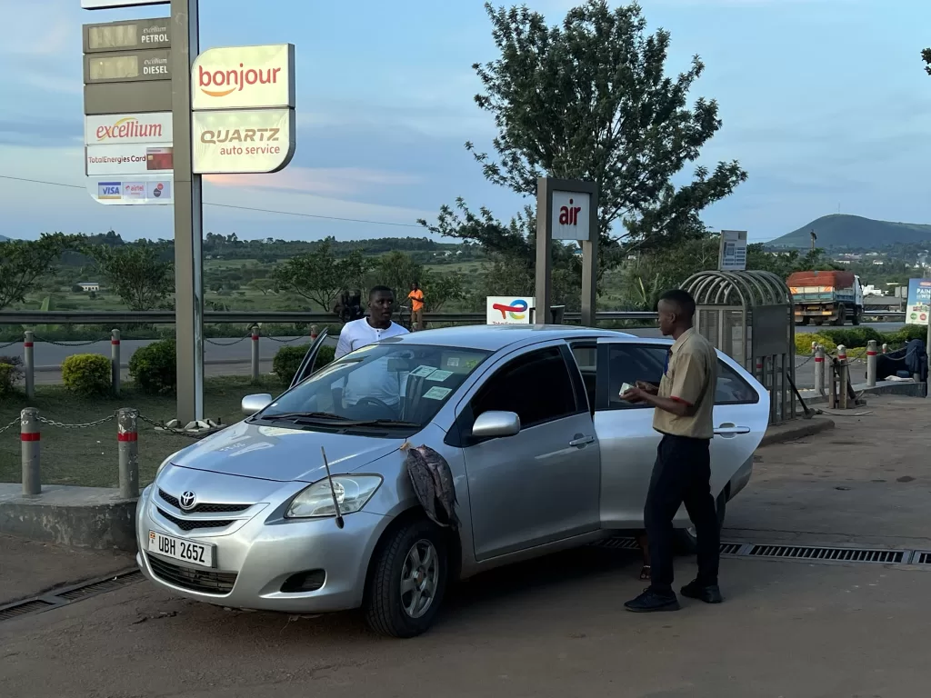 Car with fish drying on the hood of a car at a Total gas station in rural Uganda while the driver and attendant discuss filling up the tank.